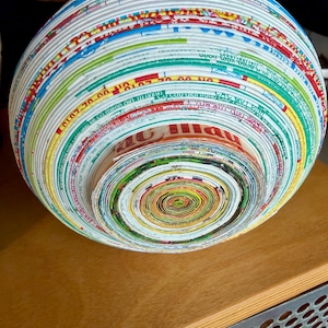 May include: A spherical bowl crafted from tightly rolled strips of colorful paper, showcasing a spiral pattern at the center. The bowl's exterior displays a vibrant array of colors, including red, blue, green, and yellow, creating a unique decorative piece.