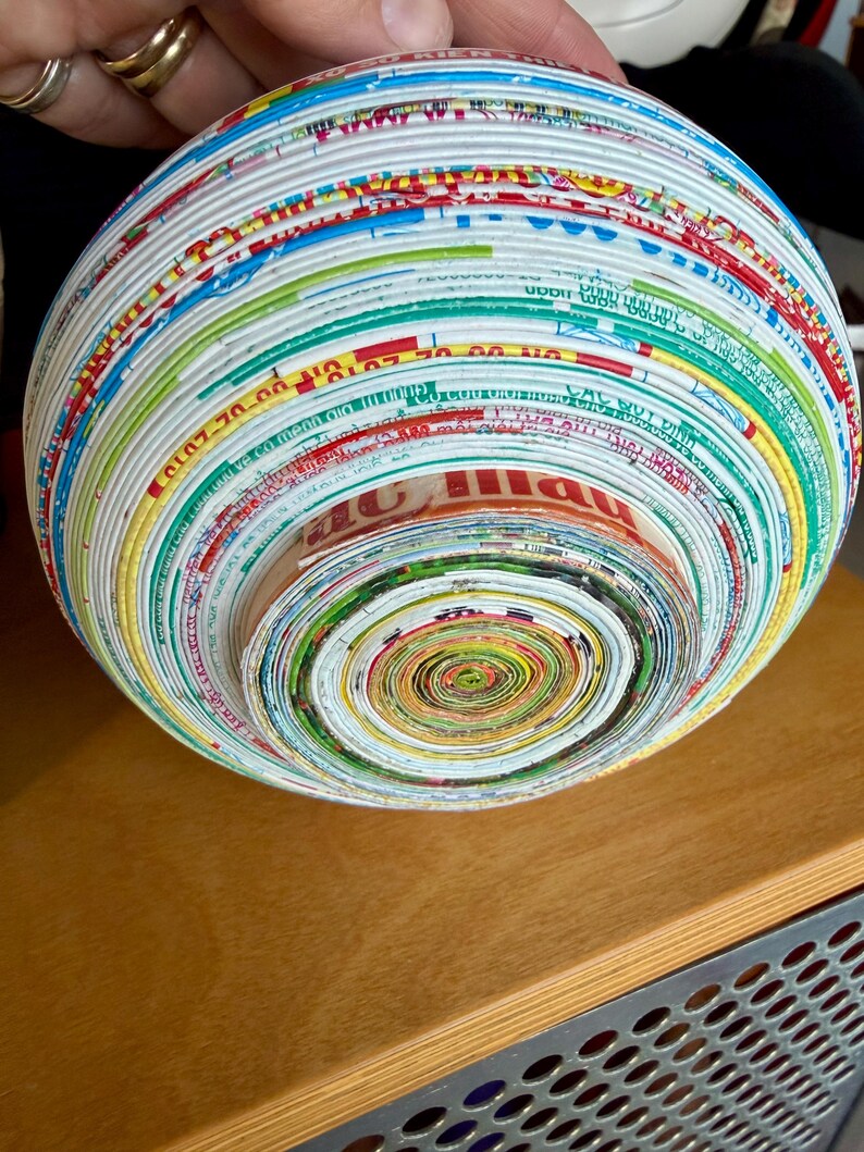 May include: A spherical bowl crafted from tightly rolled strips of colorful paper, showcasing a spiral pattern at the center. The bowl's exterior displays a vibrant array of colors, including red, blue, green, and yellow, creating a unique decorative piece.