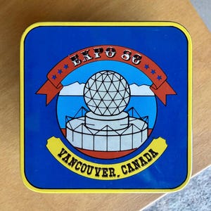 May include: A square, blue Expo 86 souvenir with a yellow border. The design features a white geodesic sphere above a white structure, with the text "EXPO 86" and "VANCOUVER, CANADA" in yellow.