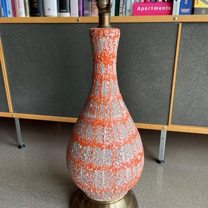 Op de afbeelding: Een moderne tafellamp uit het midden van de eeuw met een textuur, oranje en grijs patroon. De lamp heeft een messing voet en een lamphouder aan de bovenkant. De lamp is ongeveer 61 cm hoog.