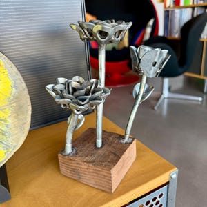 May include: A sculpture featuring three metallic flowers crafted from silver-toned hardware components, including nuts and washers, attached to thin metal stems. The stems are anchored into a square, natural-toned wooden block base.