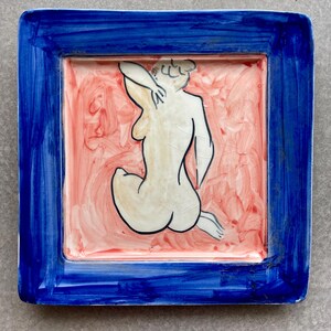 Puede incluir: Plato cuadrado de cerámica con borde azul y diseño central. El diseño presenta una figura desnuda en posición sentada, delineada en negro, sobre un fondo rosa y rojo. El plato tiene un acabado brillante.