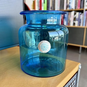 May include: A clear, blue glass jar with a wide mouth and a rounded body. The jar has a label that reads "HAND BLOWN GLASS". The jar is sitting on a wooden surface.