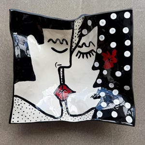 Puede incluir: Un plato cuadrado de cerámica con bordes ondulados que presenta una ilustración en blanco y negro de dos rostros besándose. El diseño incluye labios rojos, una flor roja y lunares blancos sobre fondo negro, con la firma Belkoff visible.