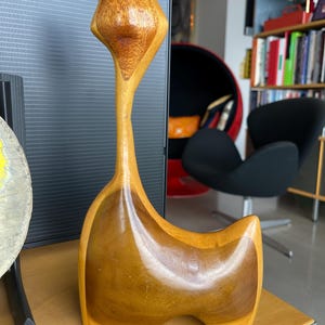 Pode incluir: Uma escultura de gato de madeira com pescoço longo e traços estilizados. O gato tem um corpo castanho claro com detalhes castanhos mais escuros na cabeça e no peito. A escultura é exibida em uma superfície de madeira.