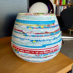 May include: A decorative, handcrafted vase with a unique design. The vase is made from layered paper strips in various colors, including blue, white, and red. The text "XỔ SỐ KIẾN THIẾT TP. HỒ CH&Iacute; MINH 10.000₫" is visible on the surface. The vase is on a wooden surface.