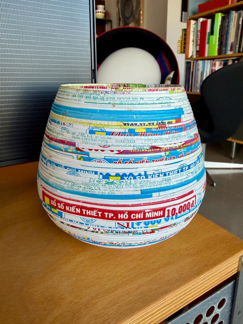 May include: A decorative, handcrafted vase with a unique design. The vase is made from layered paper strips in various colors, including blue, white, and red. The text "XỔ SỐ KIẾN THIẾT TP. HỒ CH&Iacute; MINH 10.000₫" is visible on the surface. The vase is on a wooden surface.