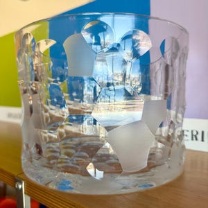 Puede incluir: Un vaso de vidrio transparente con un exterior texturizado y un patrón geométrico de formas esmeriladas. El vaso cilíndrico se encuentra sobre una superficie de madera, reflejando la luz.