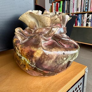 Puede incluir: Un jarrón de cerámica hecho a mano con un diseño texturizado único. El jarrón presenta una mezcla de tonos terrosos, incluyendo marrón, beige y toques de rojo y verde. La parte superior tiene una apariencia arrugada y plegada, lo que aumenta su atractivo artístico. Esta pieza decorativa es ideal para la decoración del hogar.