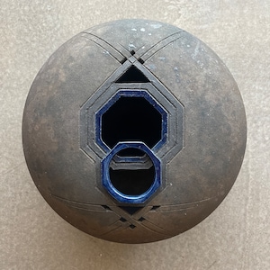 Puede incluir: Una esfera de cerámica gris oscuro con un diseño geométrico y un anillo esmaltado azul. La esfera tiene un agujero en el centro.