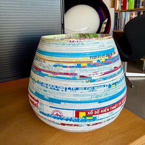 May include: A handcrafted, decorative vase with a wide, rounded body and a slightly flared rim. The vase is made from recycled paper strips, featuring a collage of colorful text and graphics in blue, white, red, and yellow. It sits on a wooden surface.