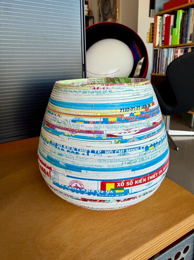 May include: A handcrafted, decorative vase with a wide, rounded body and a slightly flared rim. The vase is made from recycled paper strips, featuring a collage of colorful text and graphics in blue, white, red, and yellow. It sits on a wooden surface.