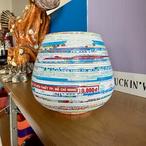 May include: A decorative, round vase made from layered paper, featuring a variety of colors and text. The vase has a wide base and tapers slightly towards the top. The text includes "SO KIEN THIET TP. HO CHI MINH 10.000d".