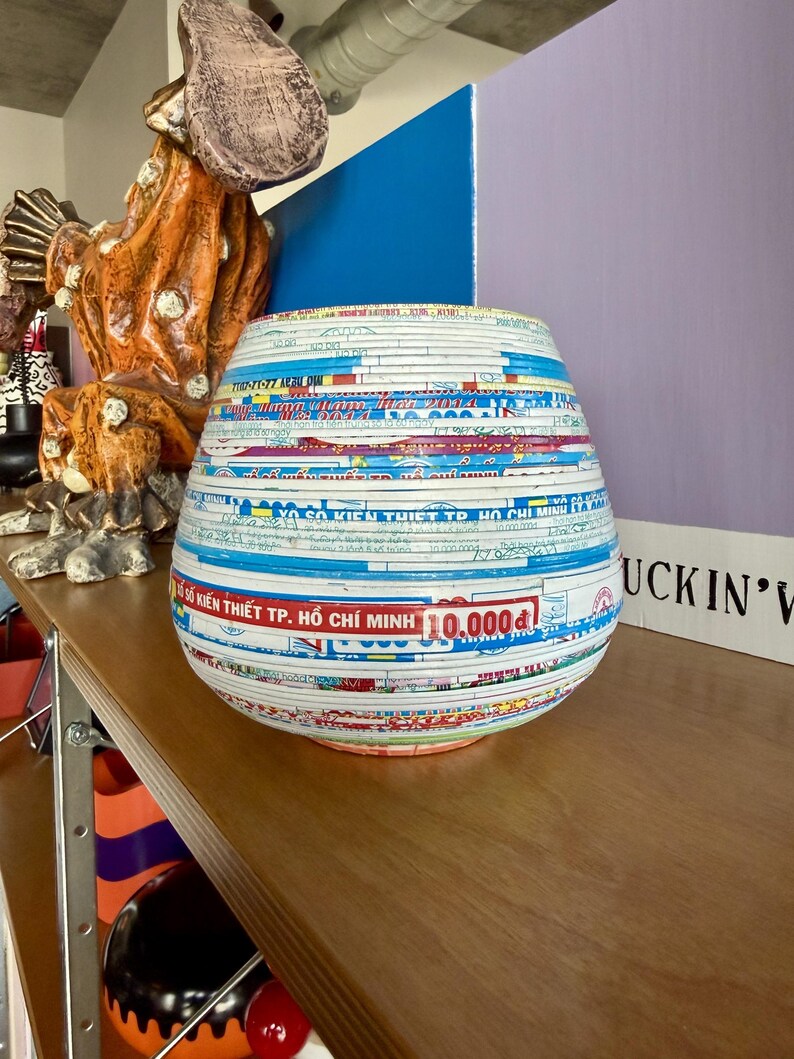 May include: A decorative, round vase made from layered paper, featuring a variety of colors and text. The vase has a wide base and tapers slightly towards the top. The text includes "SO KIEN THIET TP. HO CHI MINH 10.000d".