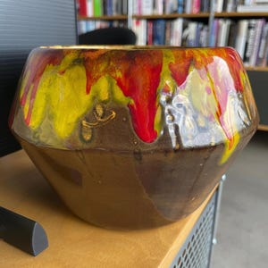 May include: A brown ceramic bowl with a unique design. The top rim features a vibrant blend of yellow, red, and gold dripping down the sides. The bowl has a geometric shape and a glossy finish, perfect for home decor.