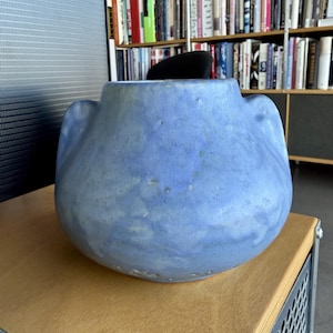 May include: A large, rounded, blue ceramic vase with two side handles. The vase has a textured surface and sits on a light-colored wooden surface. The background includes a bookshelf filled with books.