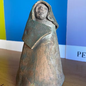 May include: A ceramic sculpture of a person wearing a hooded cloak. The sculpture is painted in shades of brown, green, and grey.