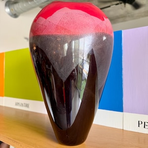 Può includere: Un vaso decorativo alto con finitura lucida. La sezione superiore è di un rosso vibrante, che sfuma in una fascia rosa strutturata, per poi passare a una base marrone scuro. Il vaso è esposto su una mensola di legno.