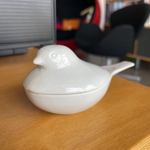 May include: A white ceramic bird-shaped container with a lid. The container is smooth and glossy, with a detailed bird design. The bird has a small tail and is sitting on a wooden surface. The container is likely used for storage or decoration.