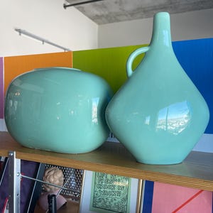 Puede incluir: Dos jarrones de cerámica de color azul aguamarina claro. El jarrón de la izquierda es redondo y el jarrón de la derecha tiene una forma más angular con un asa.