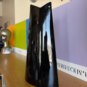 May include: A tall, glossy black vase with a distinctive V-shaped top. The vase reflects the surrounding environment, including a colorful backdrop and a silver duct. The vase is displayed on a wooden shelf.
