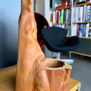 Puede incluir: Dos piezas decorativas de madera. Una es una escultura alta de forma natural con una superficie lisa de color marrón claro. La otra es un pequeño recipiente cilíndrico, también de madera, con un exterior tosco y un interior ahuecado. Ambos están sobre una superficie marrón claro.