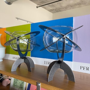 May include: Two glass vases with a black metal frame and stand. The vases are on a wooden shelf with colourful backgrounds behind them. The text on the background reads "ABSINT", "TURQUOISE", and "PERIWINKLE".