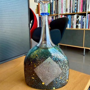 Puede incluir: Una botella de vidrio decorativa con cuello transparente y cuerpo bulboso y moteado. El cuerpo hace la transición de verde oscuro a negro con detalles amarillos y blancos. El tapón es transparente con detalles azules.