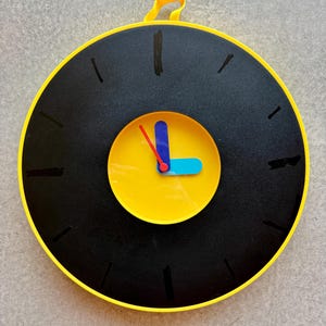 May include: A circular wall clock with a black face and yellow surround. The clock features a yellow centre with blue and red hands. Black lines mark the hours. A yellow loop is at the top for hanging. The clock's diameter is approximately 30 cm.