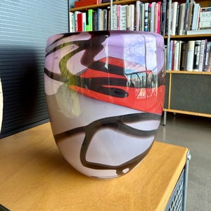 Può includere: Un grande vaso di vetro di forma ovale con un'ampia apertura. Il vaso presenta una miscela di colori lavanda, rosso e nero, con un motivo a spirale. Il vaso è appoggiato su una superficie di legno.