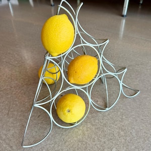 May include: A silver-colored metal wire rack with a triangular frame structure, designed to hold items in circular openings. Four bright yellow lemons are positioned within the wire frame, with one lemon in each of the three main circular sections.