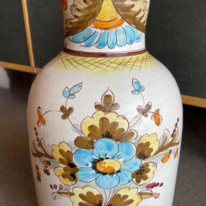 May include: A white ceramic vase with hand-painted floral designs in blue, yellow, and brown. The vase has a wide base and a narrower neck, with an orange rim. The floral pattern includes stylized flowers and leaves, creating a decorative piece.