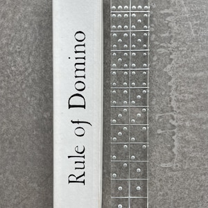 Op de afbeelding: Een doorzichtige acryl dominostenen set met witte stippen, naast een witte doos. Op de doos staat in zwarte letters "Rule of Domino". De dominostenen zijn verticaal op een grijze ondergrond geplaatst.