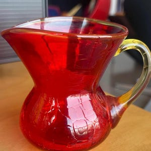 May include: A vibrant red glass pitcher with a yellow handle. The pitcher has a bulbous base and a flared top. The glass has a textured, crackled appearance. The handle is a contrasting yellow color, adding a decorative touch to the glassware.
