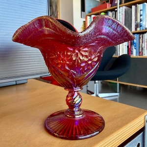 May include: A red iridescent glass bowl with a ruffled rim and a pedestal base. The bowl features a floral pattern and a twisted stem. The glass has a shiny, reflective surface, and the bowl is sitting on a wooden surface.