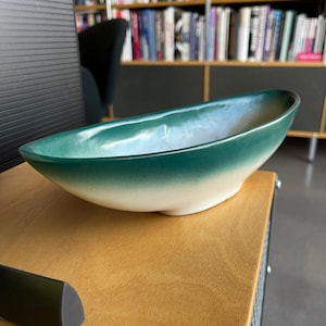 Puede incluir: Un cuenco de cerámica en forma de barco con un degradado de colores. El borde es de un verde azulado intenso, que se desvanece a un verde azulado más claro y luego a blanco en la base. El cuenco está sobre una superficie de madera.