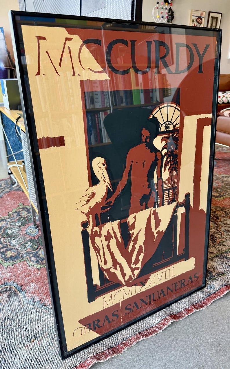 May include: Framed art print with the word "McCurdy" at the top. The artwork shows a person, a pelican, and a balcony scene. The text "OBRAS SANJUANERAS" is at the bottom. The colour scheme includes beige, brown, and black.