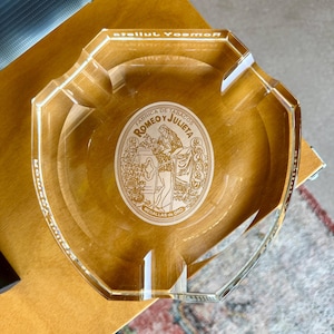 Può includere: Un posacenere in vetro trasparente con quattro poggia sigarette e forma ottagonale. Il centro presenta una stampa grafica bianca con il testo Fabrica de Tabacos Romeo y Julieta Medallas de Oro e un'illustrazione di due figure.