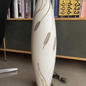 Puede incluir: Una lámpara de mesa de cerámica blanca con una base dorada y un diseño de juncos estilizados. La lámpara está en un estante de madera con libros.