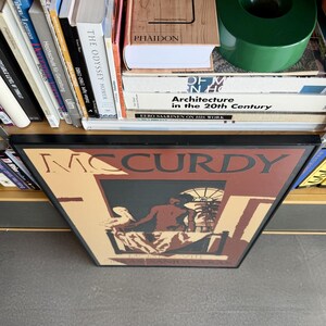 May include: A framed art print with the name "McCurdy" in large letters. The artwork shows a seated figure with a bird, set against a window and palm trees. Books and a green circular object are visible on a shelf.