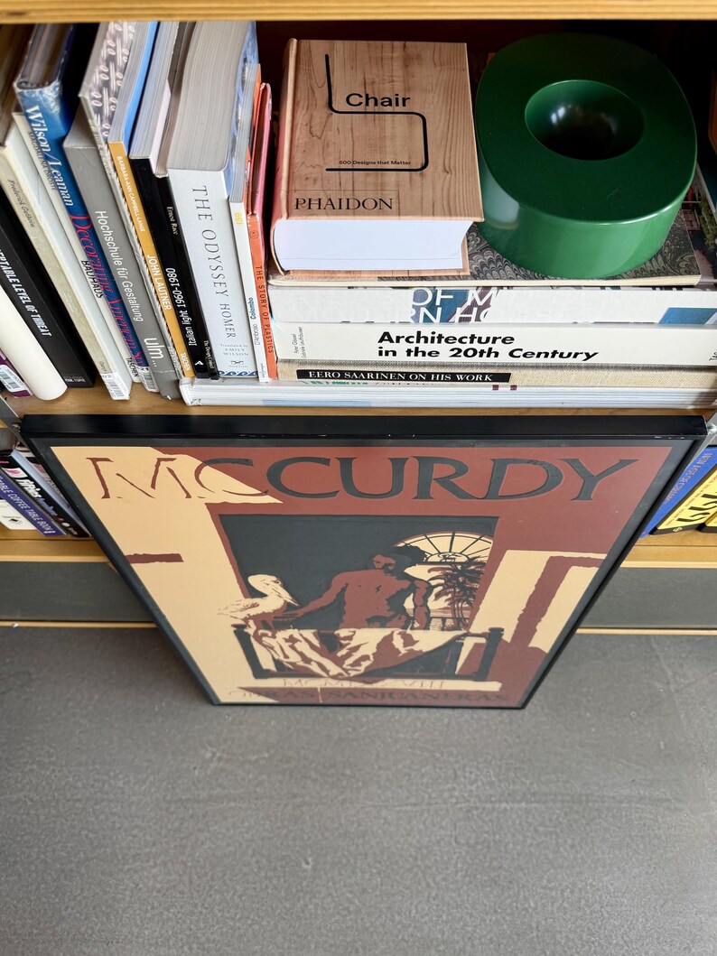 May include: A framed art print with the name "McCurdy" in large letters. The artwork shows a seated figure with a bird, set against a window and palm trees. Books and a green circular object are visible on a shelf.