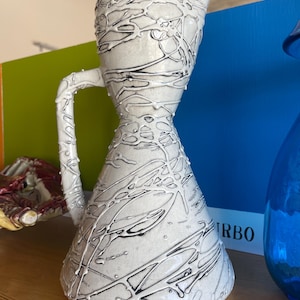 May include: A white ceramic pitcher with a black glaze design. The pitcher has a wide mouth and a narrow base. It has a handle on one side.