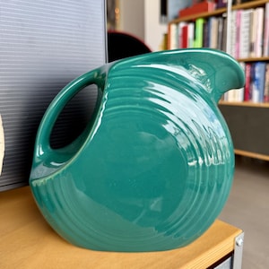 Puede incluir: Una jarra de cerámica de color verde azulado con un asa grande y curvada y un pico. La jarra tiene un cuerpo redondeado con detalles de anillos concéntricos. La jarra está sobre una superficie de madera clara.