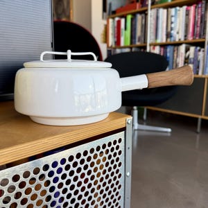 Puede incluir: Un cazo de metal esmaltado blanco con una tapa a juego que tiene un asa superior. El cazo tiene un cuerpo cilíndrico con una base escalonada y un mango horizontal único hecho de madera clara, que se extiende desde el lado del cuerpo metálico.