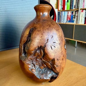 Puede incluir: Un jarrón de madera hecho a mano con un cuerpo bulboso y un cuello estrecho. El jarrón está hecho de madera marrón claro con una veta única y texturizada y nudos naturales. Es un objeto decorativo.
