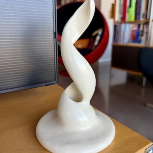 Op de afbeelding: Een crèmekleurige, abstracte sculptuur met een wervelende, langwerpige vorm. De sculptuur heeft een cirkelvormige basis en een gladde, glanzende afwerking. Het ontwerp is modern en artistiek, geschikt voor interieurdecoratie.