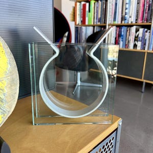 Puede incluir: Un florero de vidrio transparente con un marco exterior cuadrado y una estructura interior circular. El diseño incluye dos extensiones de vidrio que apuntan hacia arriba, creando una silueta geométrica distintiva con superficies claras.