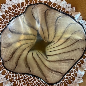 May include: A large, clear glass bowl with a swirling, abstract design in shades of brown and beige. The bowl has a black rim and a wavy, organic shape.
