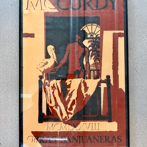 May include: Framed art print with a man, pelican, and tropical scene in orange, red, and black. The artwork includes the name "MCCURDY" at the top and text at the bottom, with a bed and window visible.
