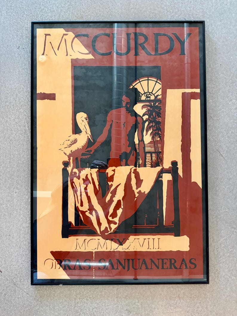 May include: Framed art print with a man, pelican, and tropical scene in orange, red, and black. The artwork includes the name "MCCURDY" at the top and text at the bottom, with a bed and window visible.
